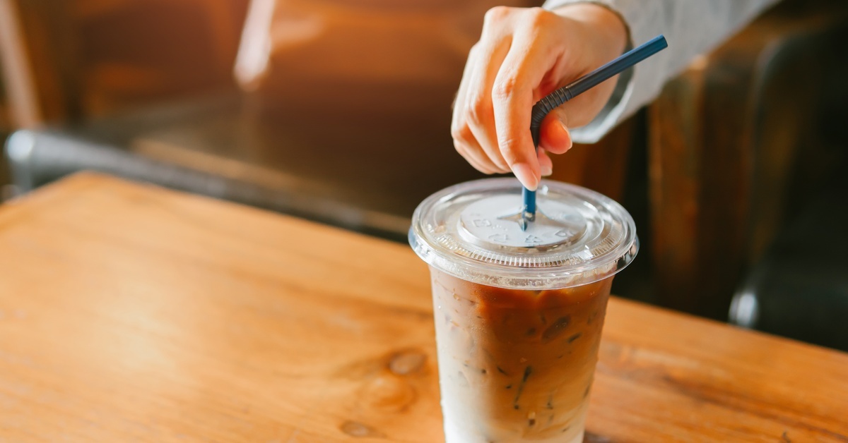 A close-up of a hand puncturing the flat lid of a clear plastic drink cup with iced coffee with a blue straw. A close-up of a hand puncturing the flat lid of a clear plastic drink cup with iced coffee with a blue straw.