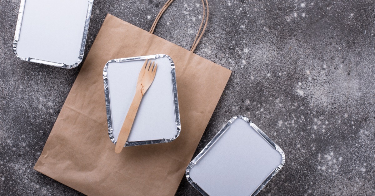 An overhead view of three aluminum-style take-out food containers. One container lies on a paper bag with a wooden spork.