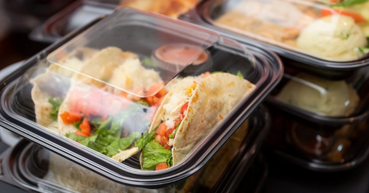 A close-up of stacks of black plastic take-out containers with clear dome lids with food inside them.