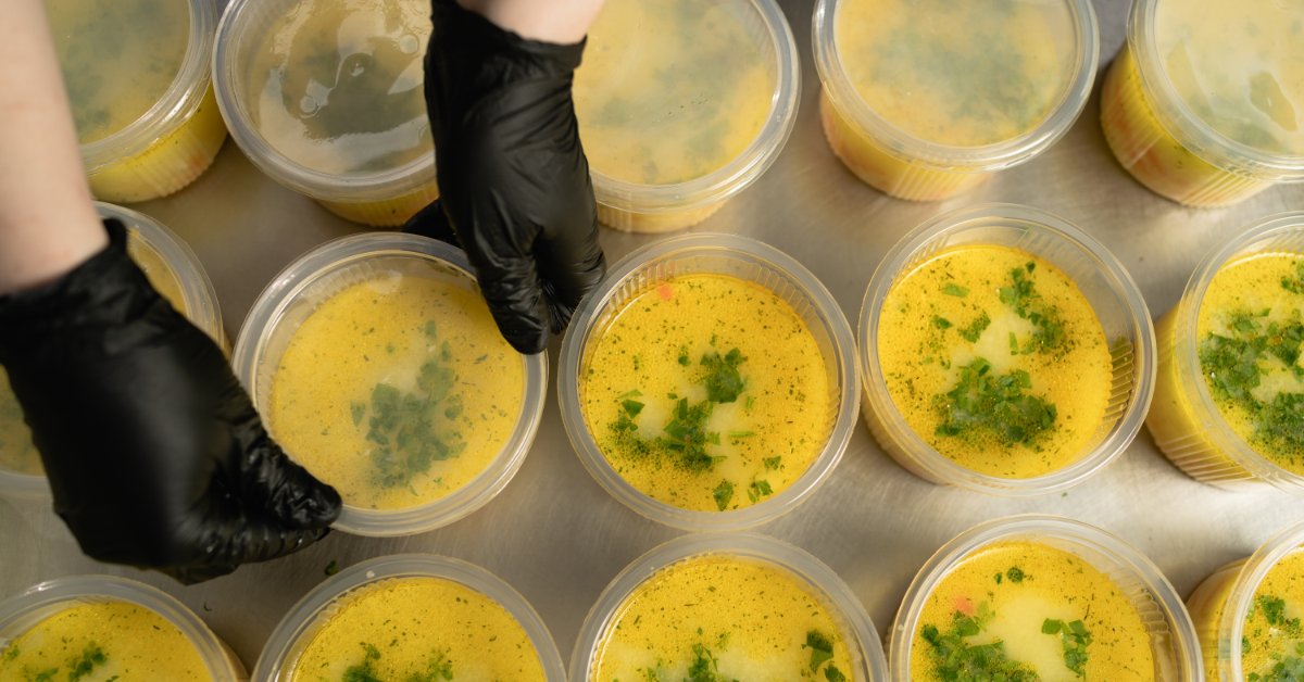 How To Choose the Right Soup Container Lid An overhead view of rows of clear to-go containers with yellow soup as a person with black gloves places lids on them.