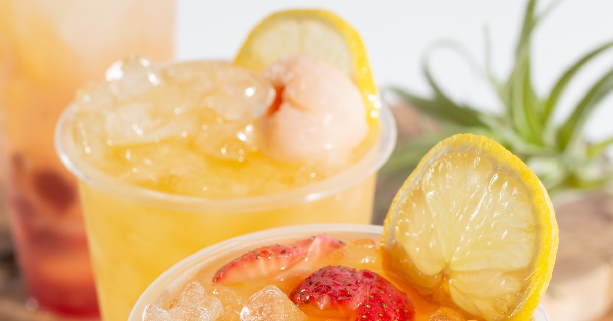 A close-up of two iced, colorful drinks with lemon and fruit garnishes in clear plastic cups without lids. A close-up of two iced, colorful drinks with lemon and fruit garnishes in clear plastic cups without lids.