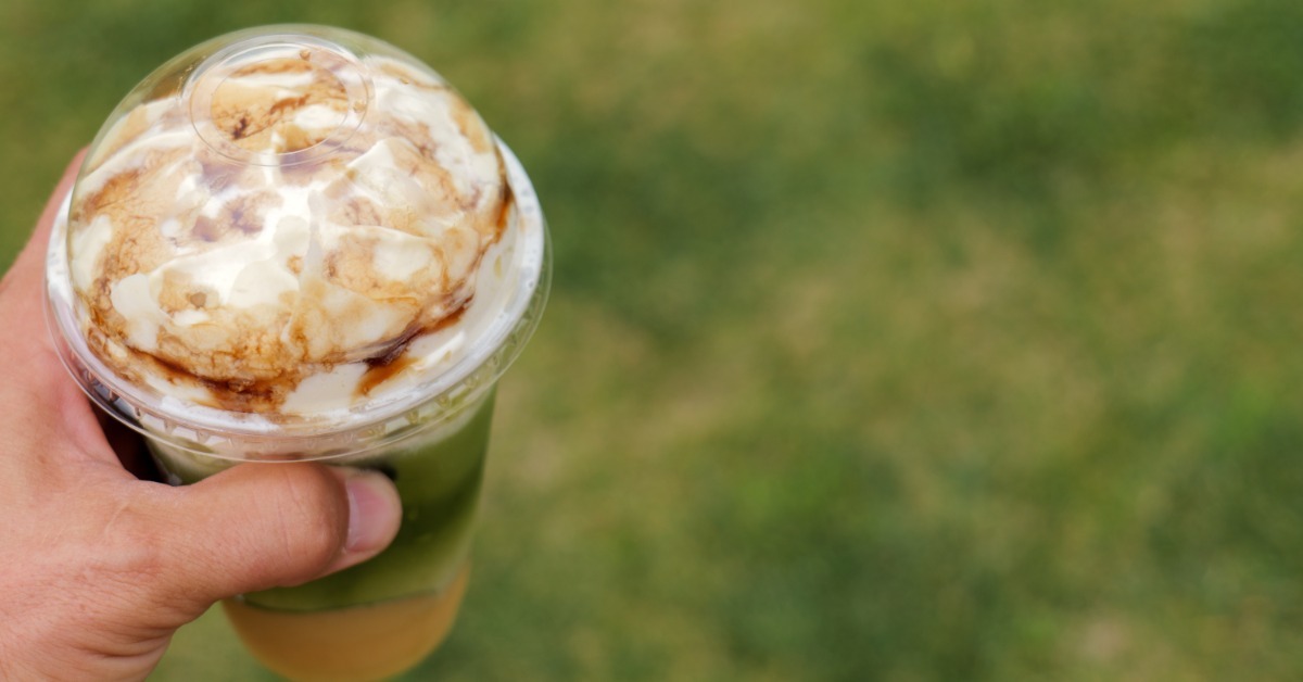 A close-up of a hand holding a clear plastic cup with a lid containing a matcha latte with a creamy foam. A close-up of a hand holding a clear plastic cup with a lid containing a matcha latte with a creamy foam.