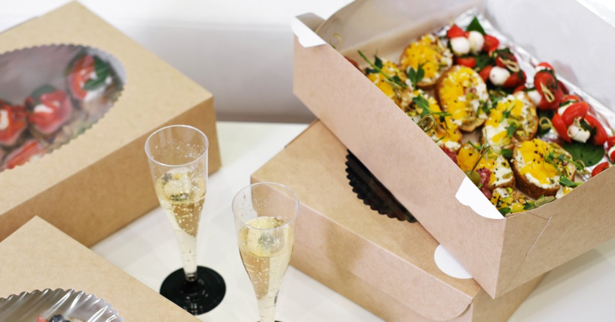 What To Expect When Ordering Custom Catering Boxes A close-up of a white table with multiple catering boxes with windows next to two glasses of sparkling wine.
