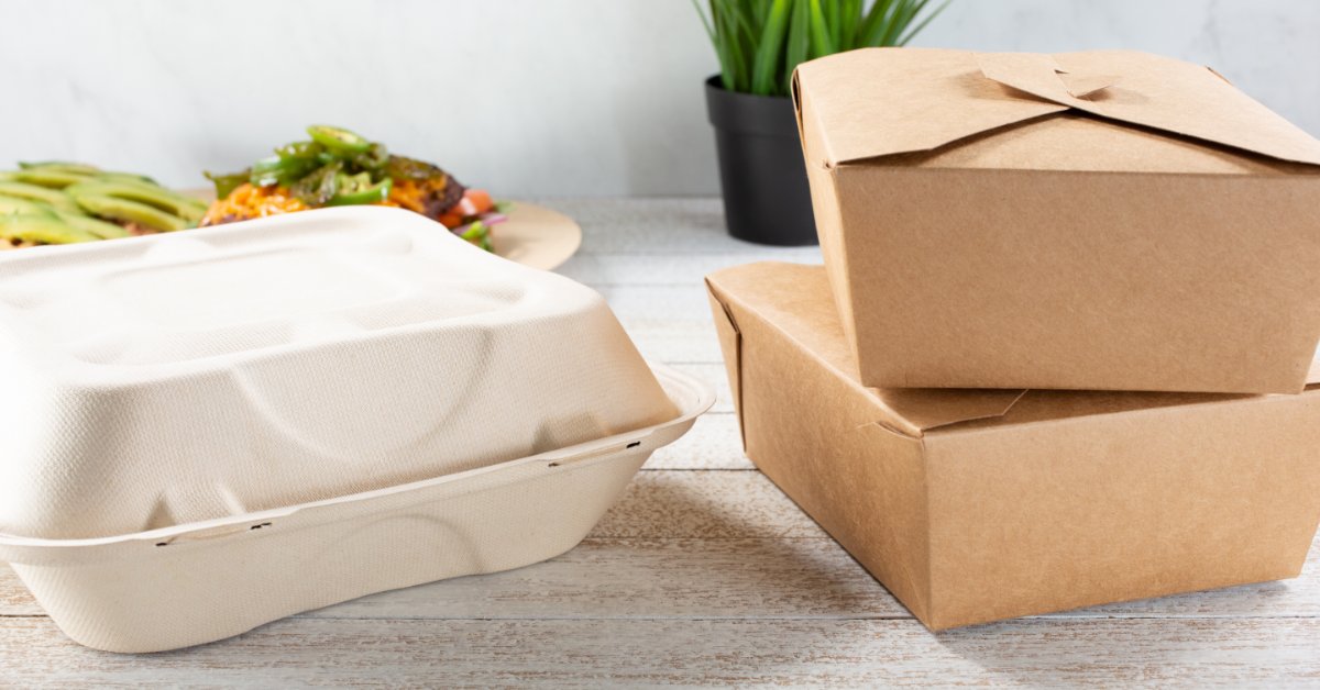 A close-up of a closed clamshell to-go food container next to two folding to-go boxes on a wooden table.