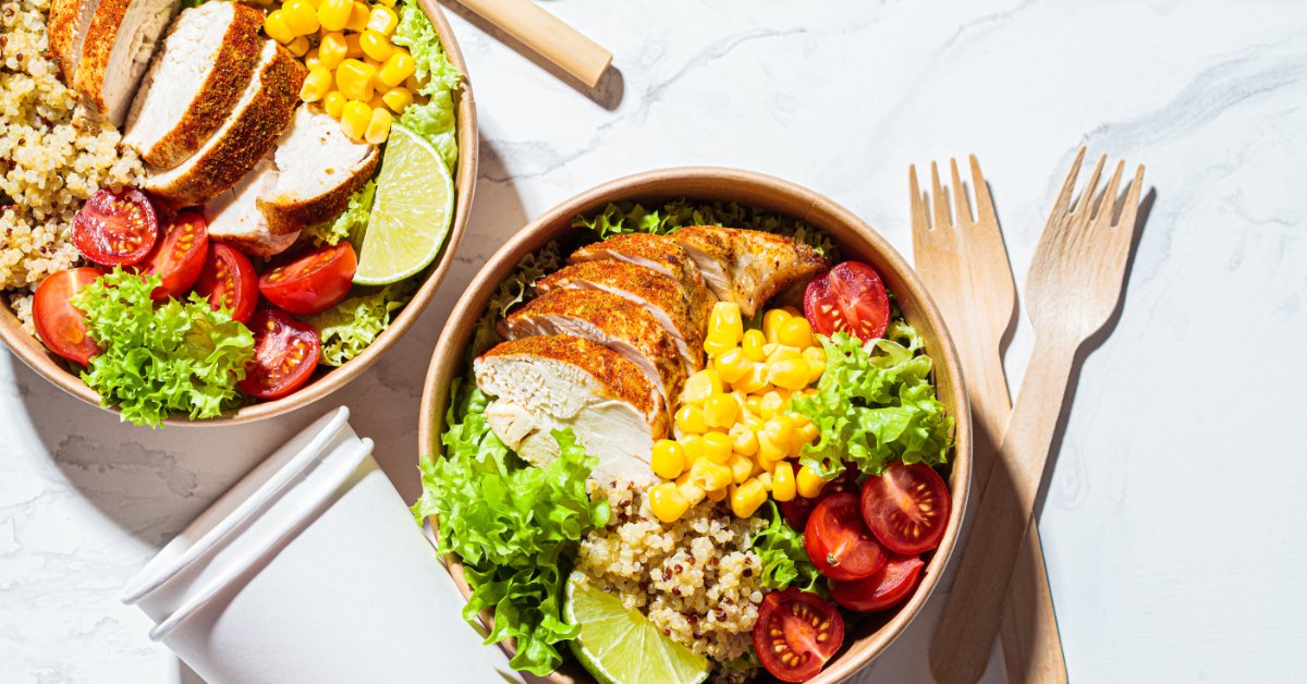 An overhead view of two quinoa salad bowls in recycled paper to-go containers next to disposable forks and cups.