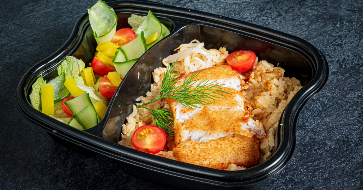 A close-up of a black plastic compartment container containing frished fish, rice, cherry tomatoes, and other vegetables.