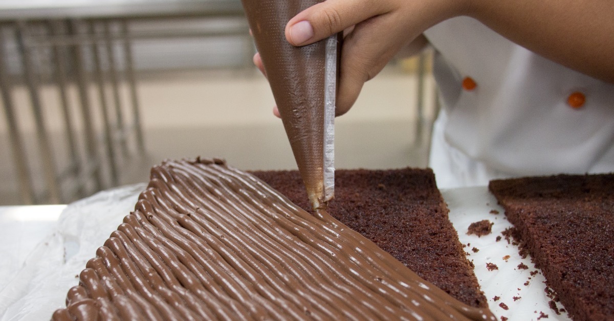 A close-up of a baker with a piping bag adding a layer of chocolate frosting to a chocolate sheet cake.