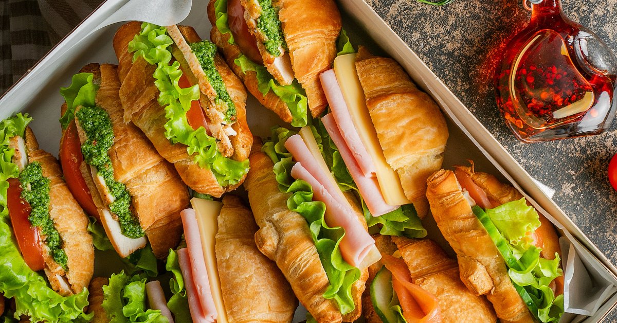 What To Expect From Heavy-Duty Catering Boxes An overhead view of a large catering box filled with croissant sandwiches on a countertop next to a bottle of oil.