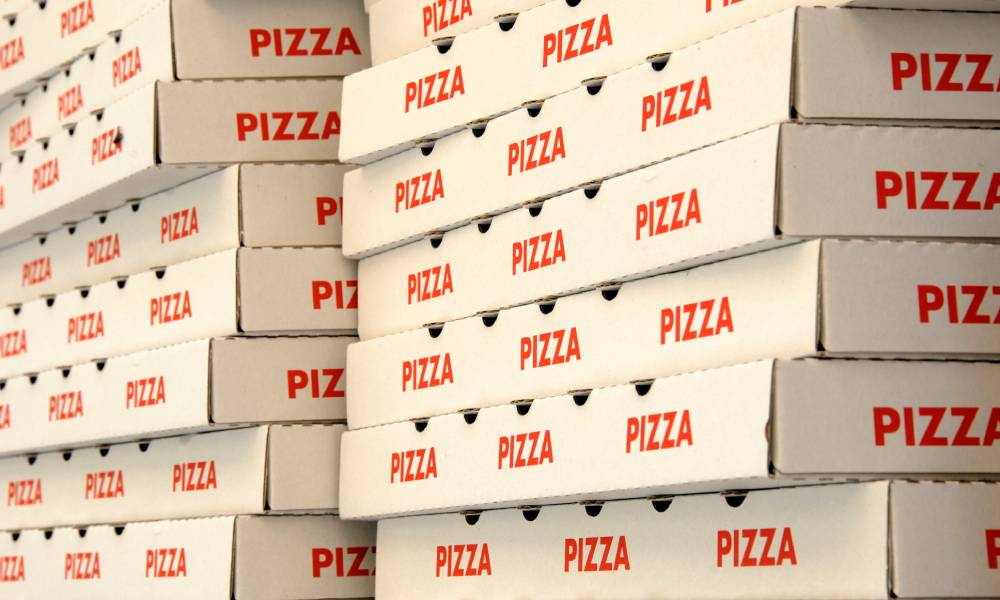 Stacks of dozens of white cardboard pizza boxes with the word 'pizza' written on them multiple times.