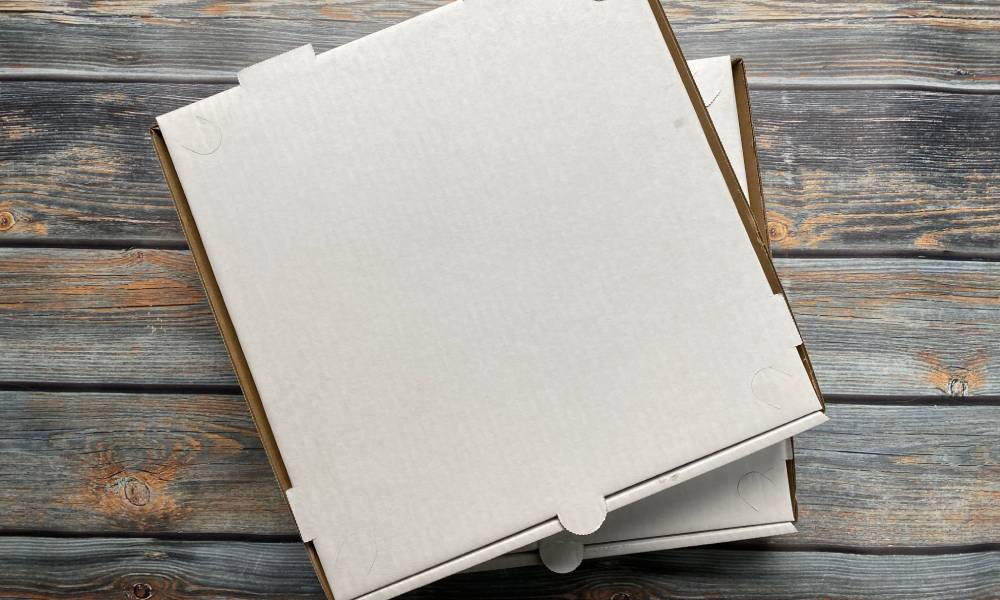 White cardboard pizza boxes stacked on top of each other on a wooden table, each with a tab on the front for easy opening.