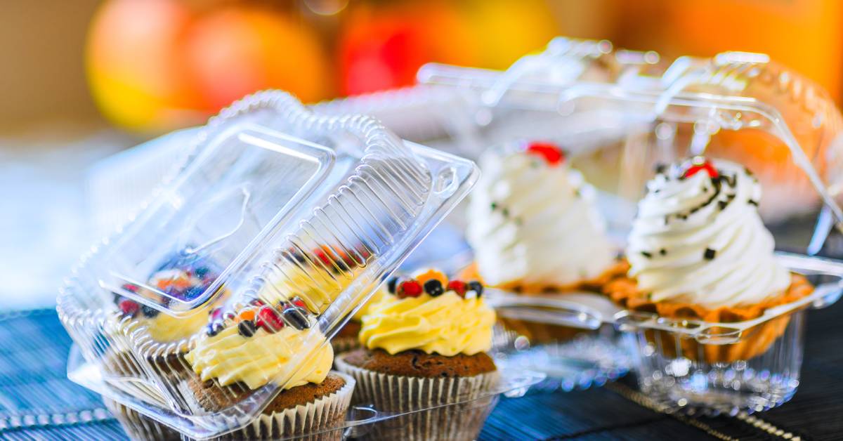 Two cupcake trays made of clear plastic full of high-end cupcakes with frosting, fruit, and other decorative elements.