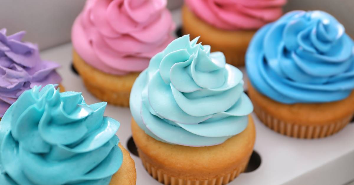 Six delicious cupcakes with pink, purple, and blue icing on white cake packaged in a white cardboard tray.