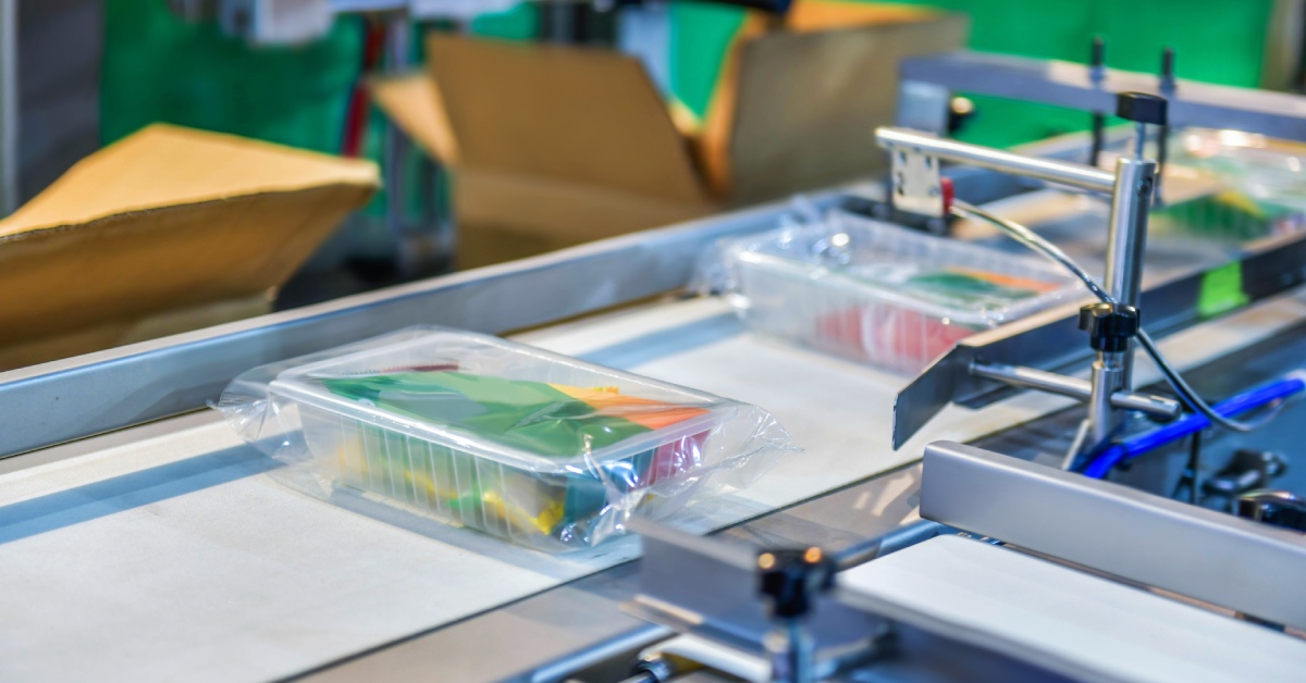 Multiple plastic boxes wrapped in shrink wrap and filled with food products on a conveyor belt in a distribution warehouse.