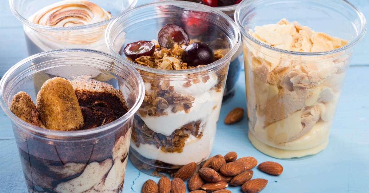 Five plastic cups filled with different dessert ice creams or parfaits on a sky-blue table with some almonds on it.