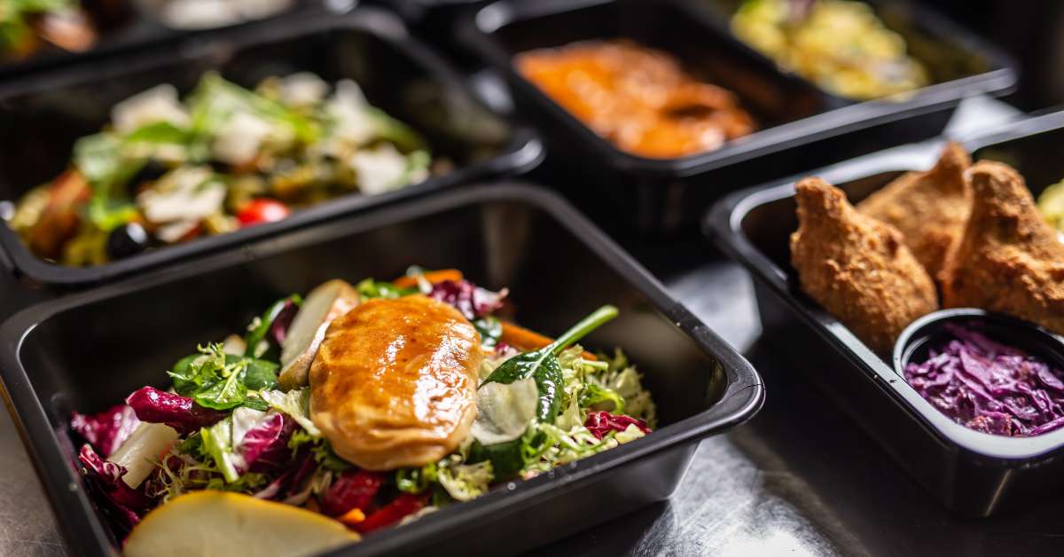 Disposable plastic food containers arranged on a metal table. The containers each hold a different meal, including a salad.