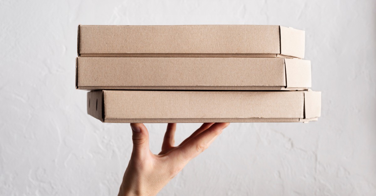 A person wearing a gray shirt with polka dots holds up three cardboard pizza boxes against a plain white background.