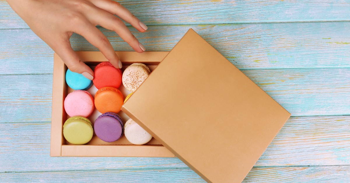 A female hand reaching toward a cardboard box of colorful macaroons resting on a painted blue wood table.