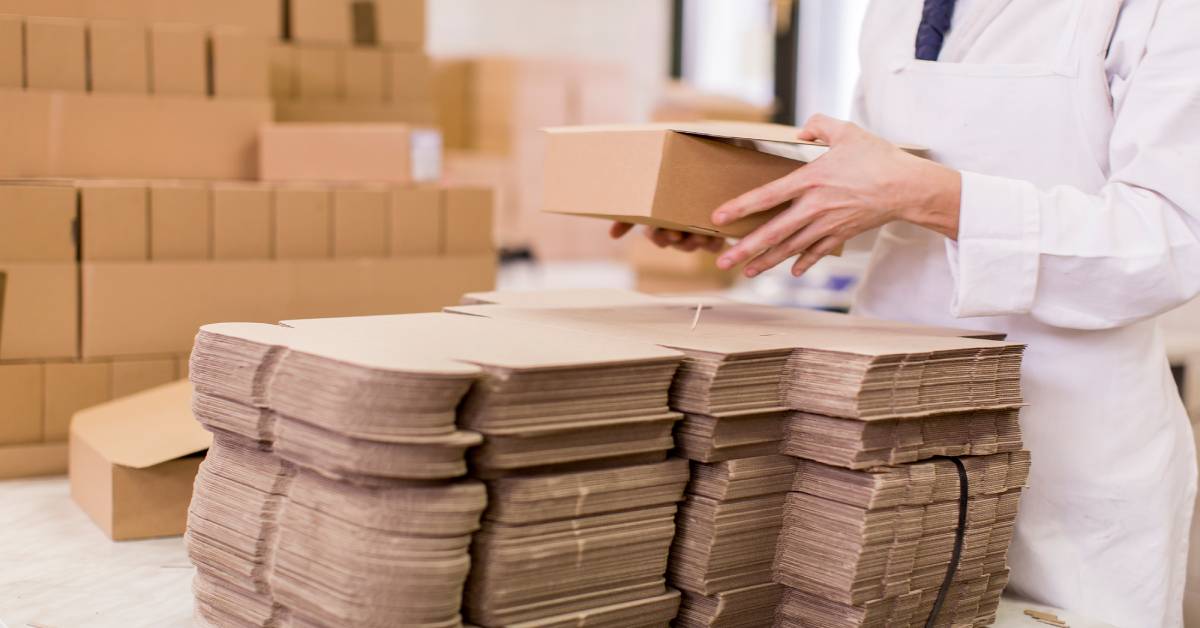 A baker in a white apron folding together a small cardboard bakery box. There is a large stack of unfolded boxes nearby.