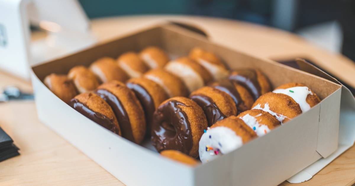 A box of assorted cake donuts with chocolate and vanilla icing and colorful sprinkles. The box is on display in a bakery.