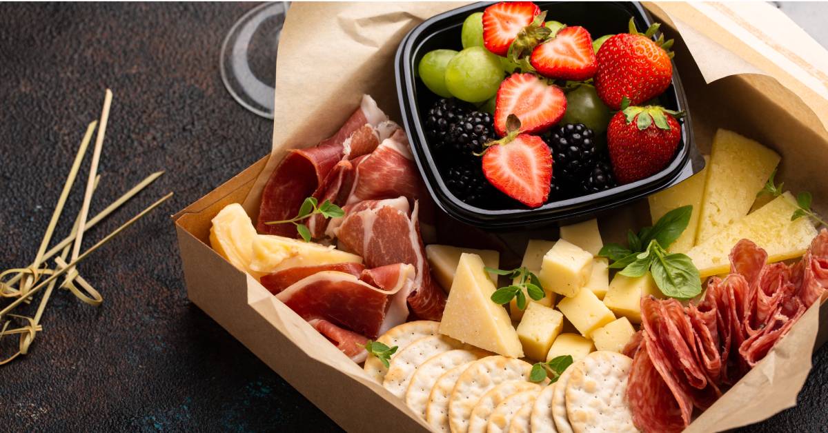 A small cardboard box filled with charcuterie food like crackers, cheese, and meat, with fruit on the side.