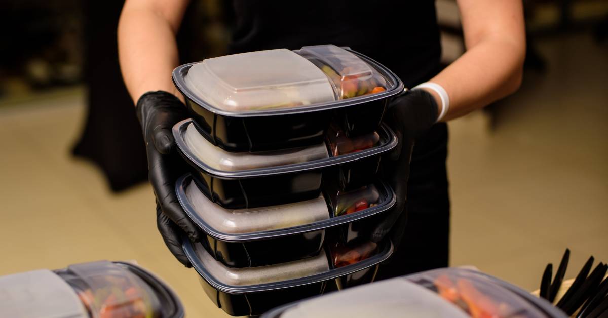 A man wearing a pair of black latex gloves delivering stacks of plastic catering containers full of hot food.