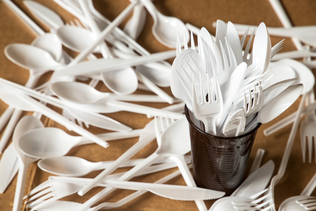 A haphazard pile of plastic forks, spoons, and knives resting on a table, some gathered in a black plastic cup.