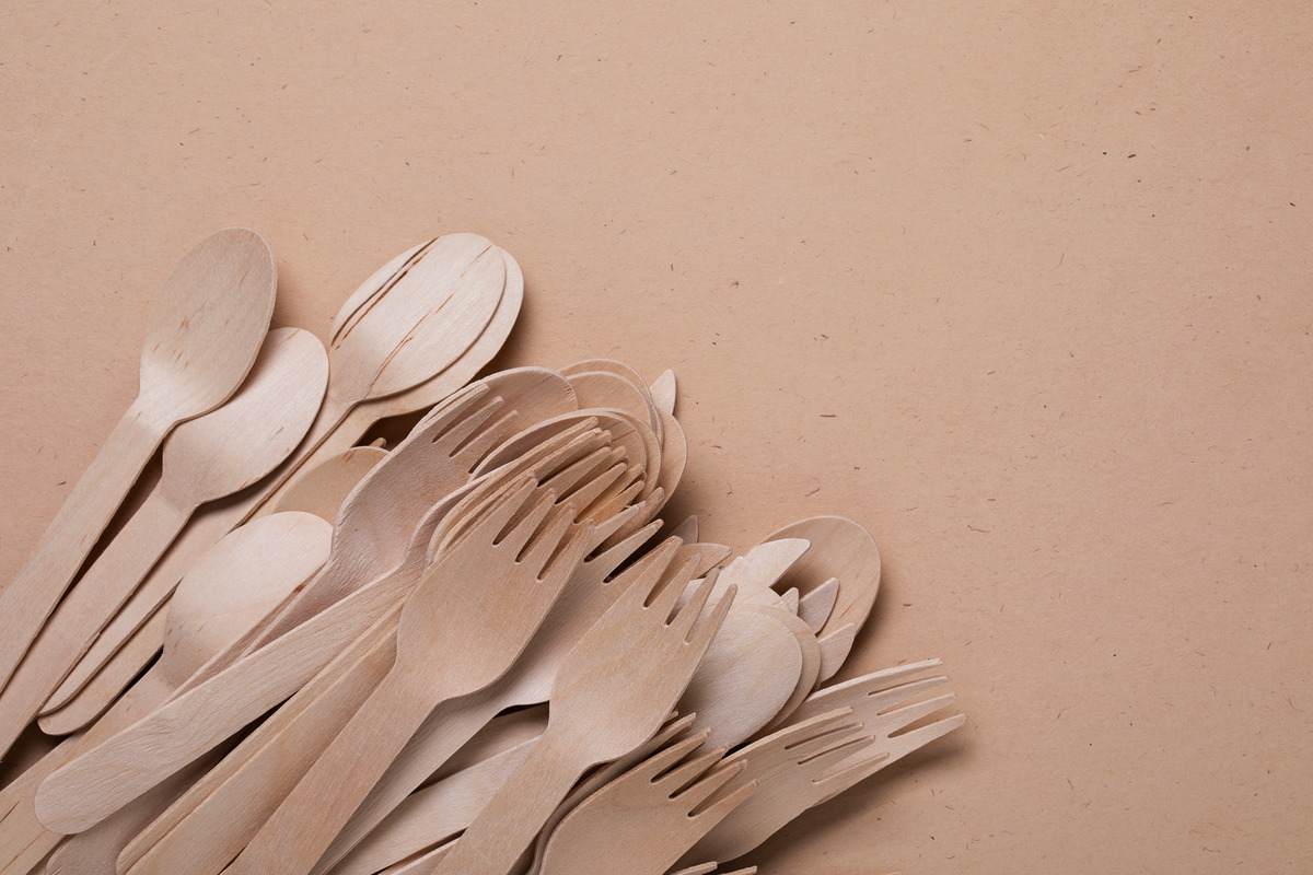 A stack of disposable and eco-friendly wooden spoons, sporks, and forks on a plain brown background.