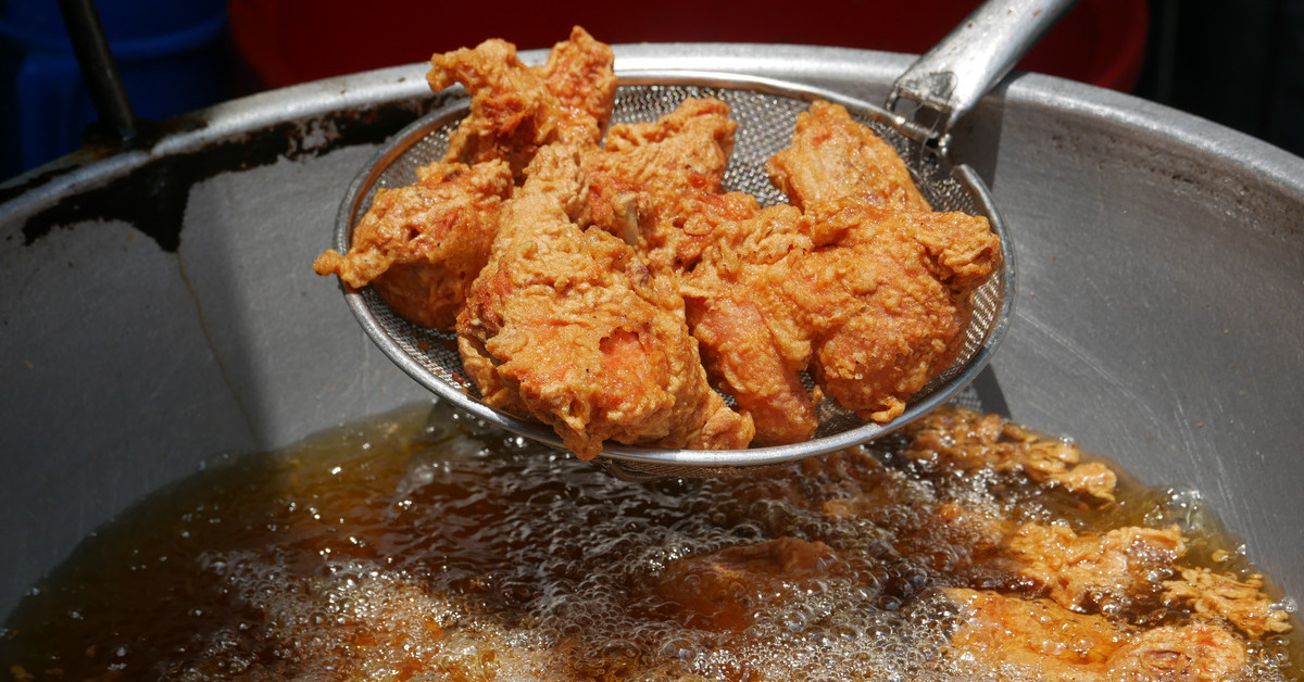 A pot of heated fry oil full of fried chicken, as someone lifts a basket of fried chicken up and out of the pot.