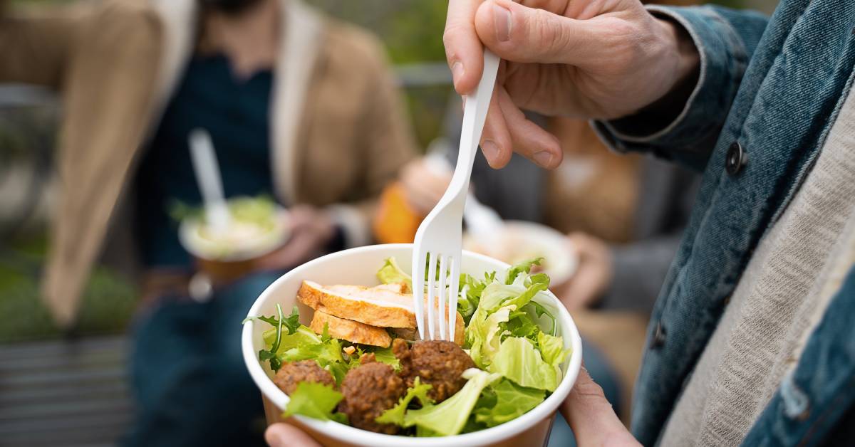 People in a park eating chicken salad out of recyclable cardboard takeout containers and using plastic forks.