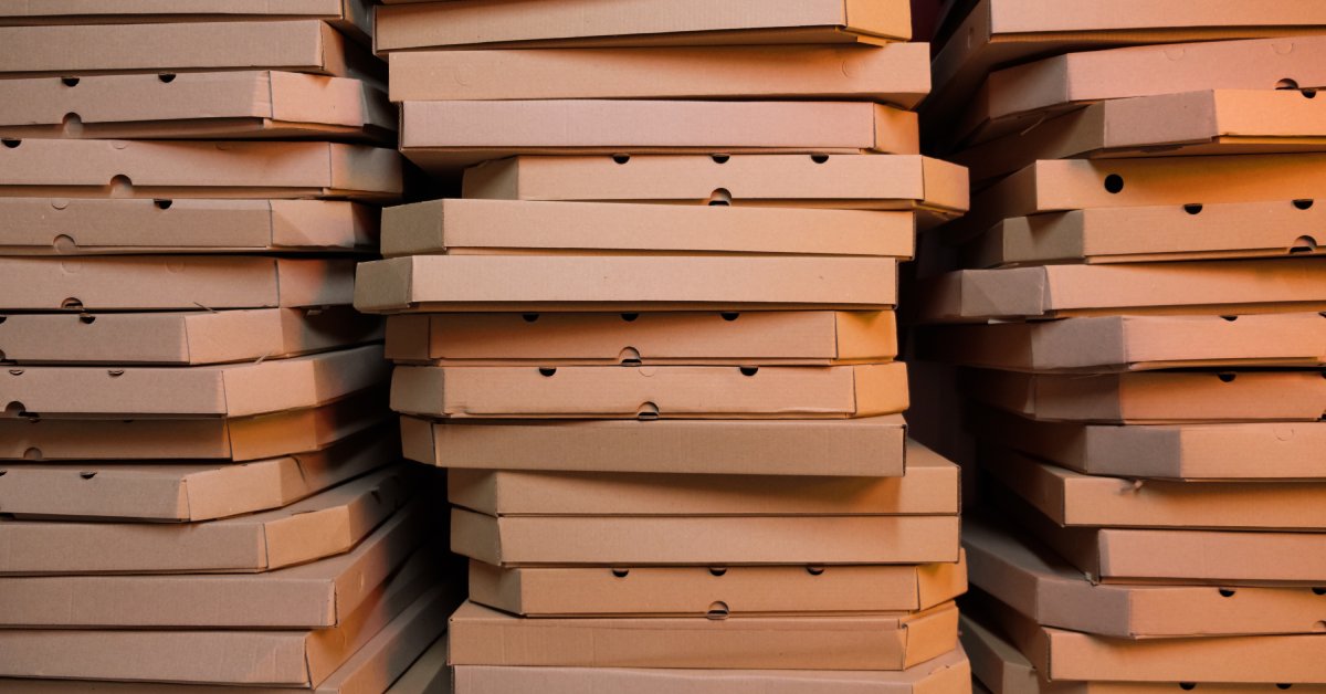 A close-up on three stacks of dozens of cardboard pizza boxes, lit from the right with a warm light.