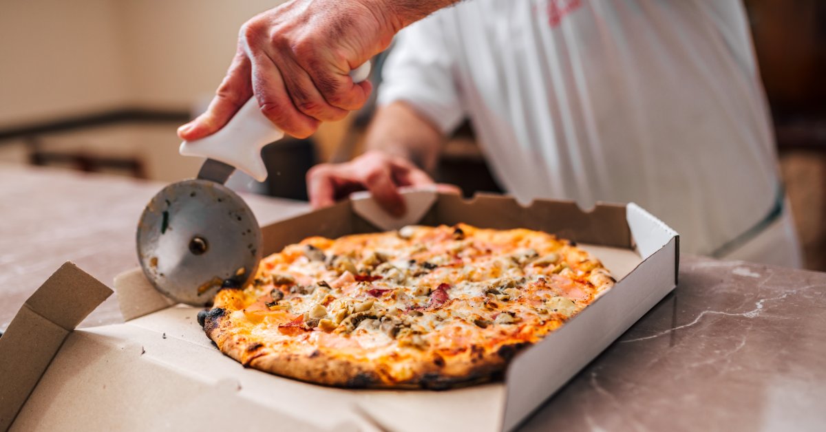 A chef cutting a circular pizza with a circular pizza cutter, the pizza is in a white cardboard box and topped with meat and vegetables.