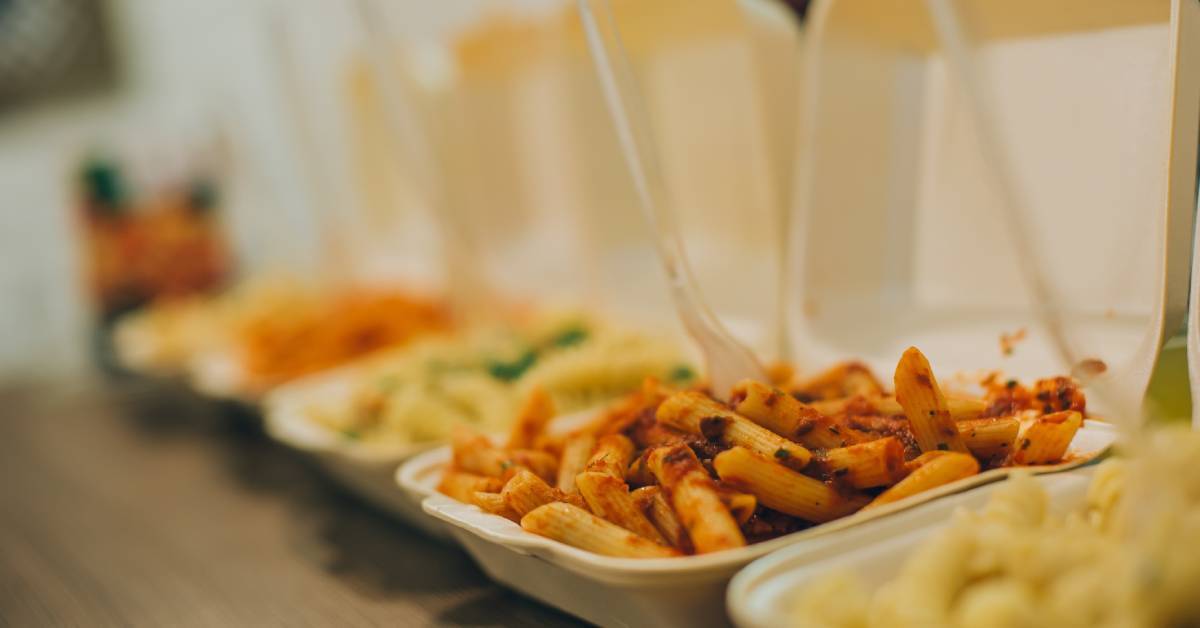 Five takeout containers with various types of pasta, including white and red sauces, each with a fork sticking out of them.