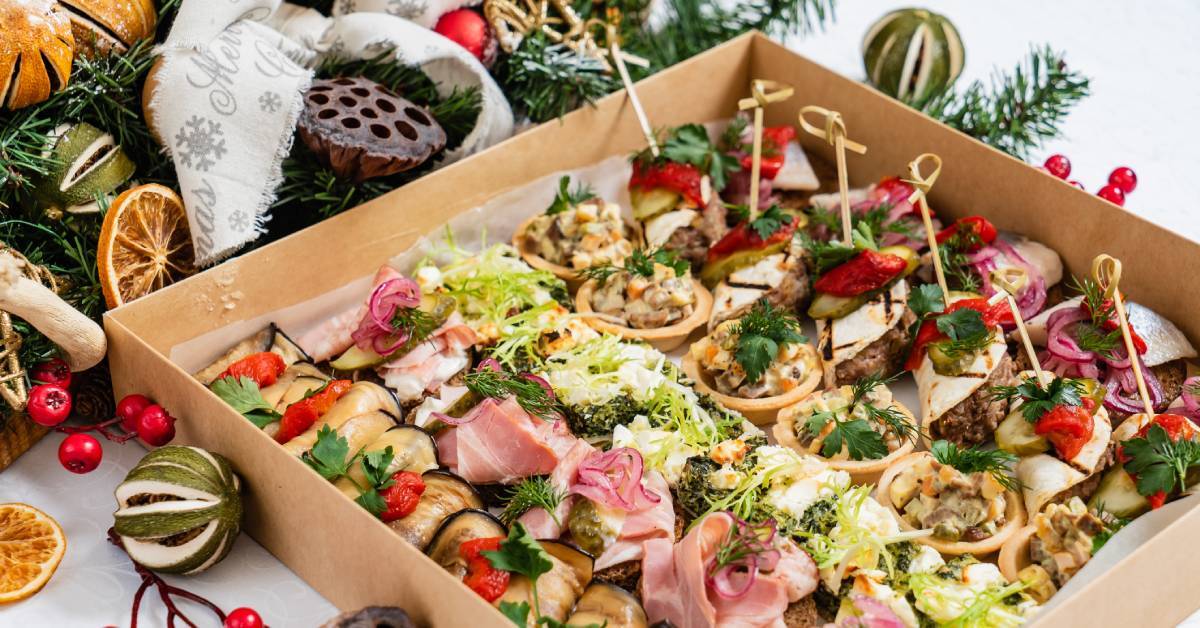 A catering grazing box on a white table surrounded by festive decorations, and filled with appetizer-like snacks.