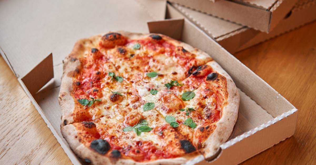 A freshly-baked pizza with mozzarella and basil in a cardboard pizza box on a wooden table with other boxes.