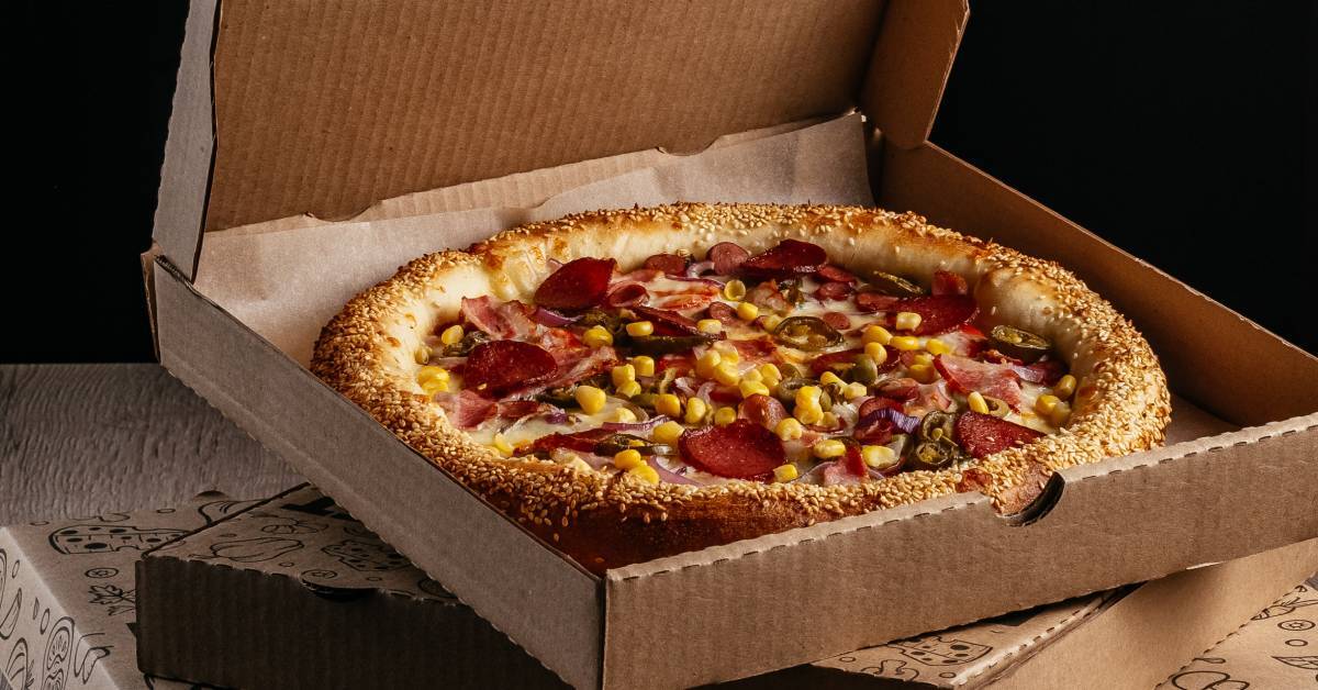 A pizza with stuffed crust and topped with meat and corn in a brown cardboard box stacked on top of other boxes.