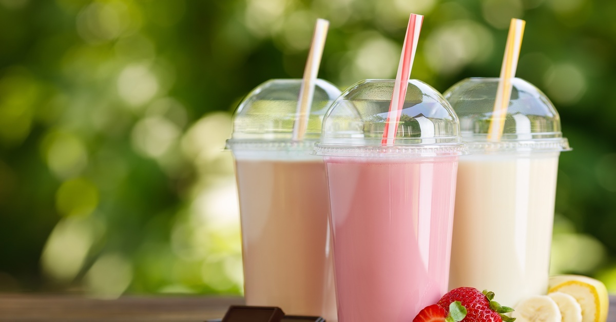 Three different smoothies in PET plastic cups with pieces of chocolate, strawberries, and bananas resting on the table with them.