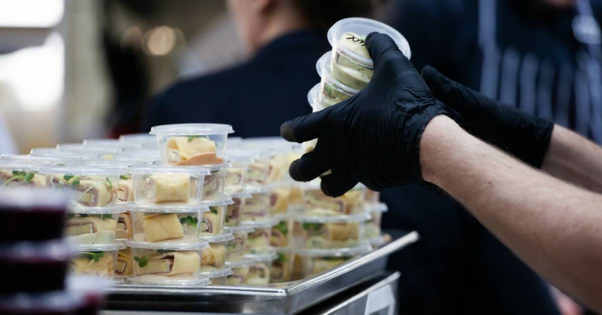 A person wearing black latex gloves stacking several single-serve appetizers in clear plastic containers on a tray.