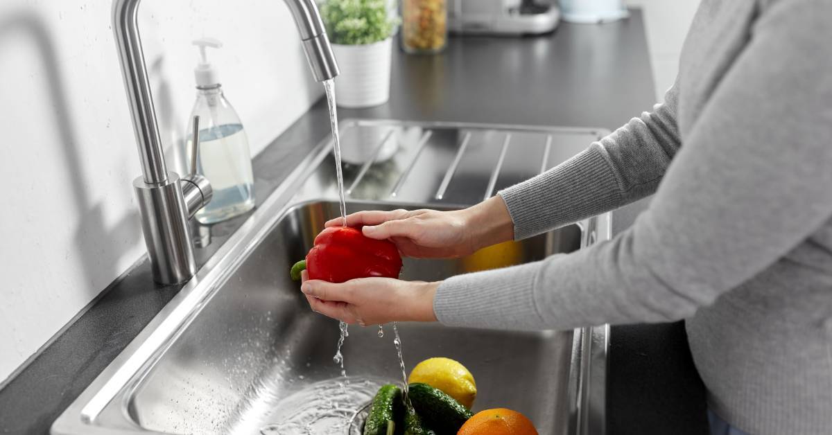 A person in a grey sweater washing several fruits and vegetables, including peppers and citruses, in a stainless steel sink.