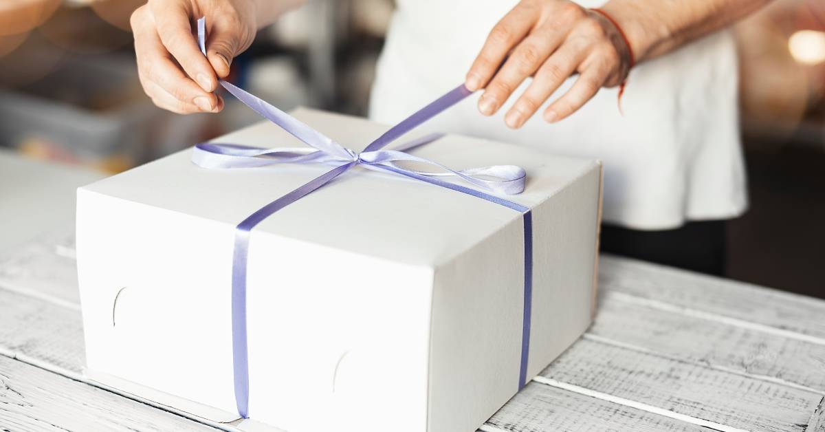 A baker in a bakery ties a purple ribbon on a white cake box that rests on a wooden table painted white.