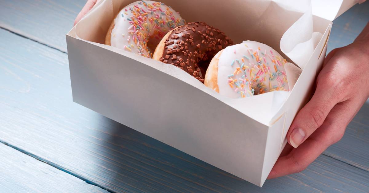 A woman holding out a white bakery box of three donuts with sprinkles above a wooden table.