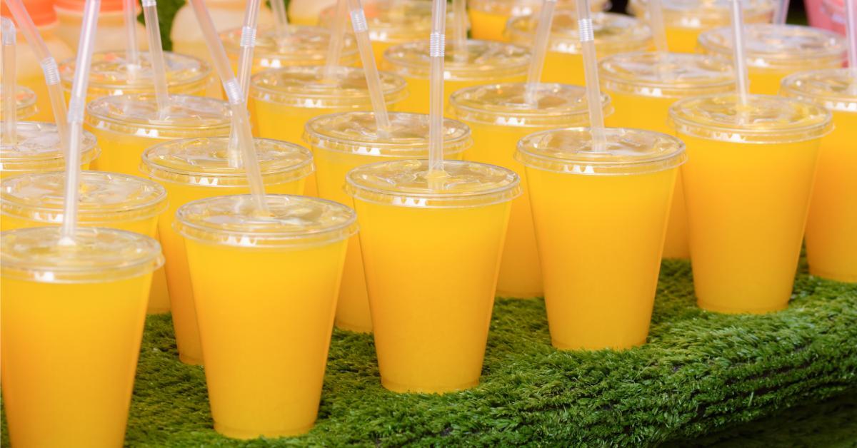 Over a dozen PET plastic cups with plastic lids and straws full of orange juice and arranged on a counter covered in green turf.