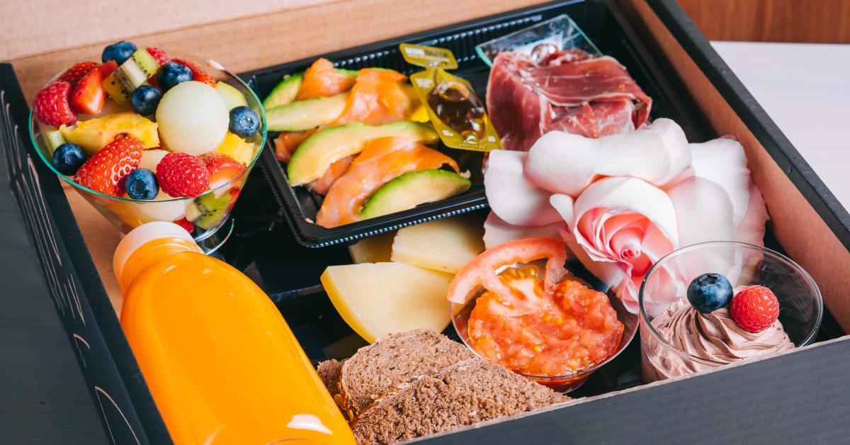 A cardboard box contains multiple food items, including fruit, ham, bread, a beverage, and a dessert.