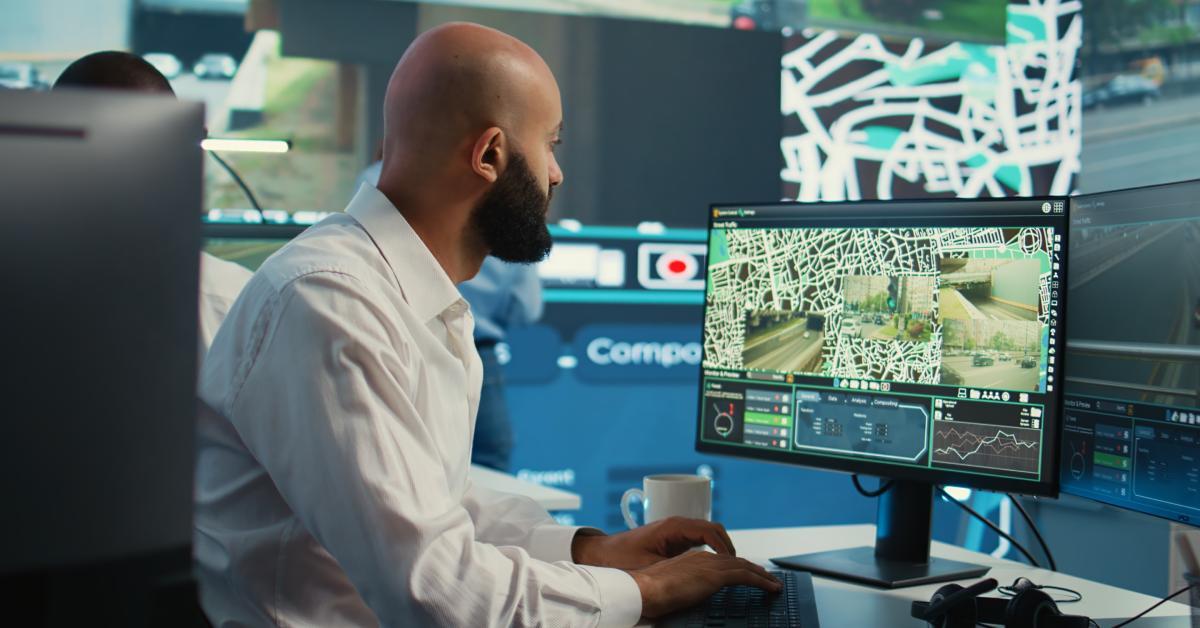 A man sits at a desk while monitoring truck shipments on his computer screen. He wears a white shirt.