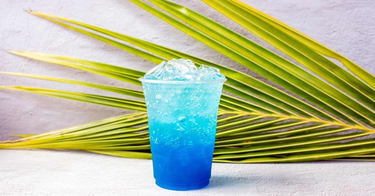 A plastic cup holds a blue drink and ice inside it. It sits in front of a background featuring coconut leaves.