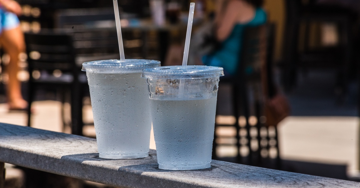 Two carbonated drinks sit in clear plastic cups with lids and straws. The two cups sit on a metal railing.
