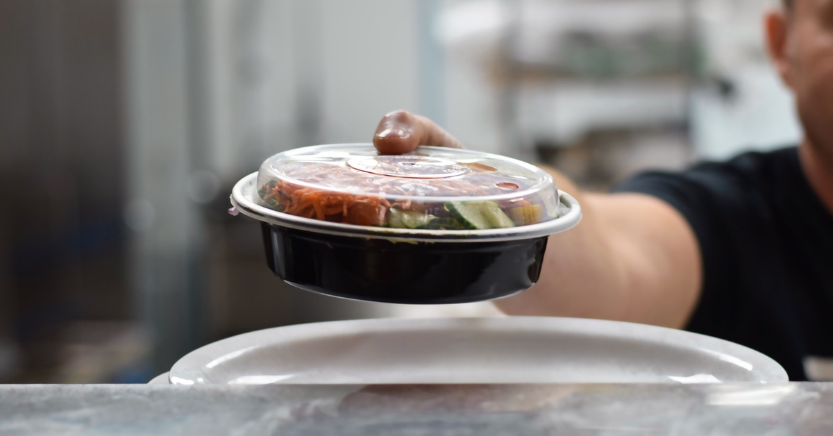 A restaurant employee holds out a plastic food container filled with a salad. The container has a clear lid.