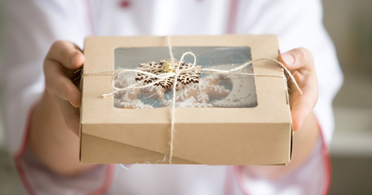 A chef's hands hold a bakery box tied shut with a piece of string. Frosted cookies are visible through the box's window.