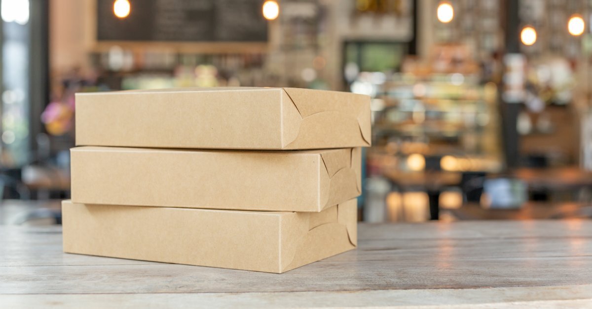 Three rectangular brown boxes sit in a pile on a counter. A store's environment is visible in the background with lights.
