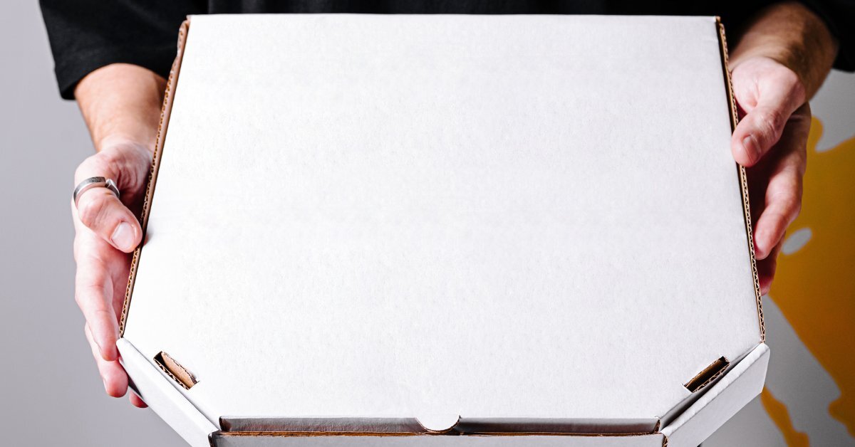 A close-up of a person in a black shirt, holding up a plain white pizza box with both hands.
