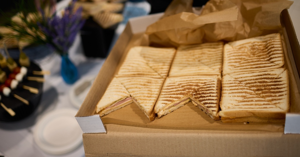 A catering box sits open on a table at an event, surrounded by other items. The box features grilled sandwiches.